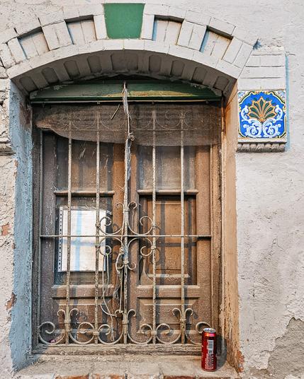 A shuttered window in poor condition but with a brightly coloured tile beside it. There is a red drinks can on the windowsill.
