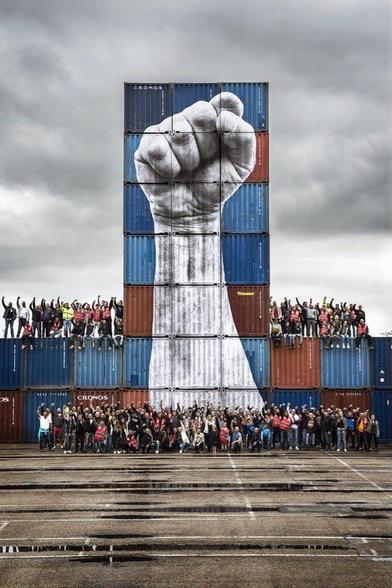 6 big containers on top og each other. A big fist is painted on them. 

100 of people standing around with raised fists.