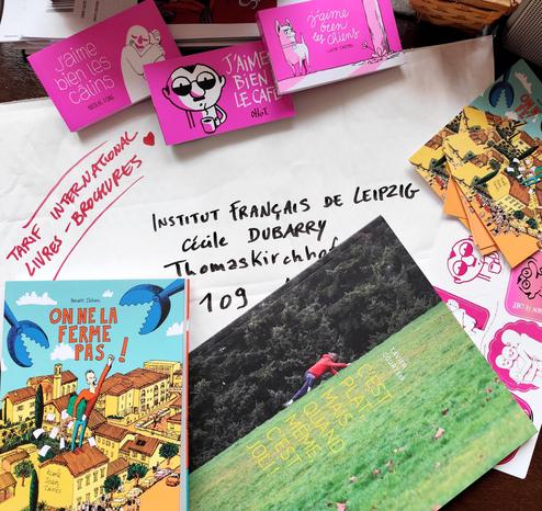 Photo de 2 livres (On ne la ferme pas ! De B. Jahan et C'est plat, mais quand même, c'est joli, de X. Courteix) et 3 flipbooks (J'aime bien les câlins, de N. Fong, J'aime ien le café, d'Otto T., j'aime bien les chiens, de L. Castel) sur une enveloppe matelassée adressée à l'institut français de Lepipzig, avec mention du tarif livres brochures - et un petit coeur.