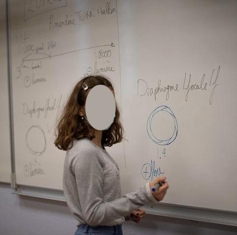 Une collégienne est devant le tableau, sur lequel est écrit les éléments clés pour apprendre l photographie en mode manuel : diaphragme, obturateur...