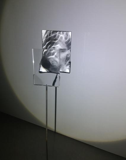 What looks like a clear, rectangular piece of glass on a stand, projecting an image of the face of a young woman on the wall behind it.