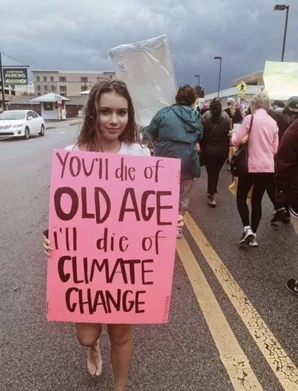 A young woman with long, wavy brown hair stands barefoot on a wet street during a protest march, holding a bright pink sign with bold black lettering. The sign reads: 'You'll die of OLD AGE, I'll die of CLIMATE CHANGE.' Behind her, a diverse group of protesters, some wearing rain jackets and carrying signs, march down the street under a cloudy, stormy sky. The scene conveys urgency and concern over climate change, with the wet pavement and dark clouds adding to the dramatic atmosphere.
