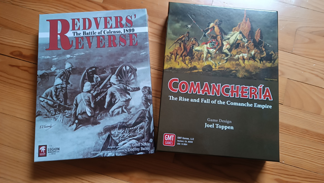 Das Bild zeigt zwei Brettspiele, die nebeneinander auf einem Holzboden liegen. Links befindet sich das Spiel "Redvers' Reverse: The Battle of Colenso, 1899" mit einem Cover, das eine monochrome Illustration von Soldaten und einer Kanone zeigt. Rechts liegt das Spiel "Comanchería: The Rise and Fall of the Comanche Empire", dessen Cover eine farbenfrohe Darstellung von Comanchen zu Pferd in einer bergigen Landschaft zeigt. Die Titel beider Spiele sind in auffälligen Schriftarten hervorgehoben.