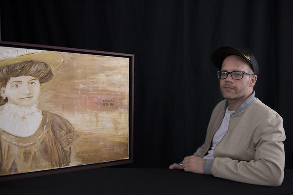 Links im Bild findet sich ein Gemälde von Emilie Rau in gelb-braunen Farben. Der Rahmen ist braun mit einem dunklen Rotstich. Ihr Geburts- und Todesjahr ist angegeben: 1891-9141.
Rechts auf dem Foto ist Andreas Hechler mit einem hellen Blouzon, einer schwarzen Brille und einem dunklen Basecap. Er blickt in die Kamera.
Emilie Rau und Andreas Hechler sitzen sich zugewandt gegenüber.
Der Hintergrund ist komplett schwarz.
Das Foto ist von Björn Hoffmann 2022 aufgenommen worden. Das Gemälde ist von Andreas Hechlers Onkel 2019 gemalt worden.