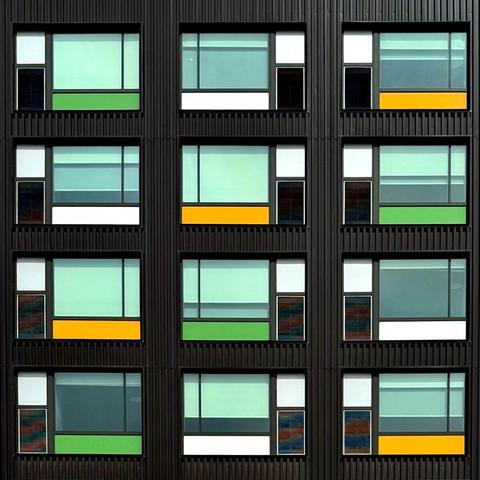 Windows on the side of a building. Overall, the building is black, but the windows have decorative panels in yellow, green, and white. The windows have additional variations based on how much the window shades are up or down. It sorrrrrt of looks like a Mondrian, but with a different color palette from the usual primary colors.