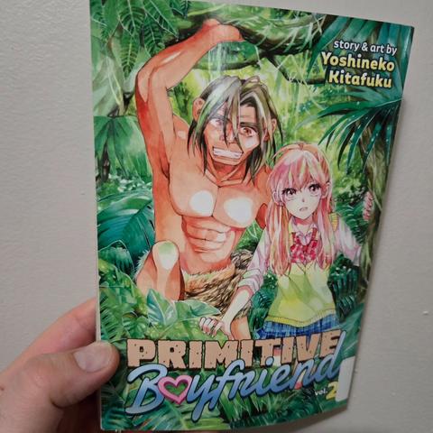 Photograph of a white person's hand holding Volume 2 of Primitive Boyfriend against a light colored wall. The cover shows a teenage girl in a Japanese school uniform in a very lush jungle next to a nearly naked ancient man who is pulling back some branches.