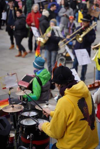 Unser Schlagzeuger von hinten, wie er gerade auf sein mobiles Drumset haut. Im Hintergrund weitere Musiker:innen an Akkordeon, Horn, Posaune und Trompete, sowie etwas Publikum.