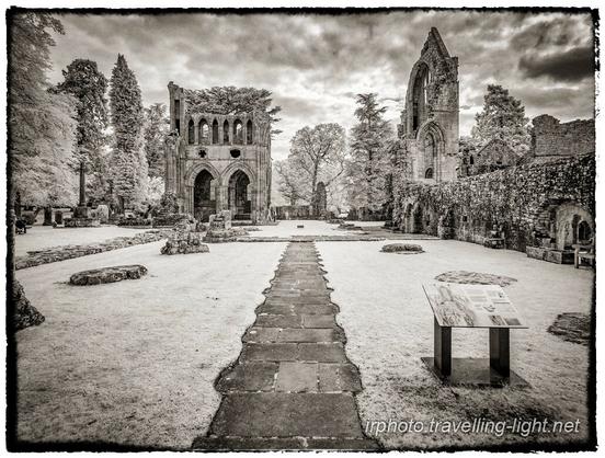 A toned black and white infrared photo of a central paved path running through the ruins of a large church.