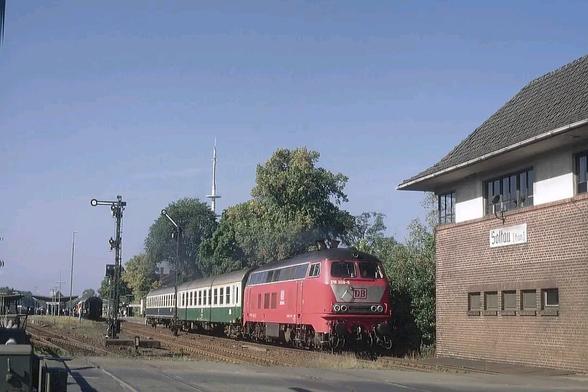 Eine rote Diesellokomotive der Baureihe 218 der Deutschen Bahn verlässt mit einem Regionalzug den Bahnhof Soltau. Der Zug besteht aus zwei grüß-weißen Reisezugwagen, darunter ein ehemaliger Wagen der Deutschen Reichsbahn. Rechts im Bild steht das Stellwerk aus rotem Backstein mit dem Schild „Soltau (Han)“. Links sind klassische Formsignale und weitere Gleisanlagen zu sehen. Im Hintergrund ragt ein markanter Sendeturm in den blauen Himmel. Die Szene wirkt typisch für den regionalen Bahnverkehr der 1990er Jahre.