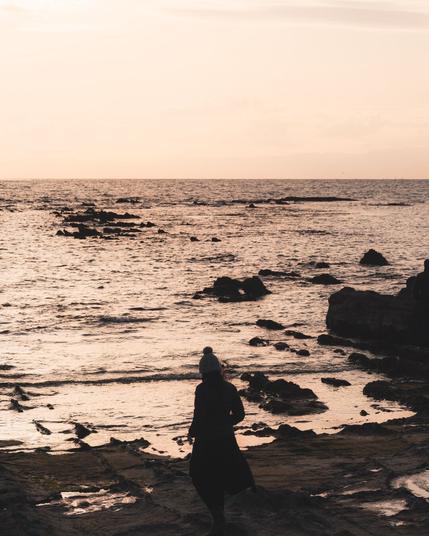 The image shows a person standing by the seashore at sunset. The ocean is visible, with waves gently hitting the rocky shoreline. The sky has a warm, golden hue typical of a sunset. The person appears to be wearing a coat and a hat, possibly due to cool weather. The overall atmosphere is calm and serene.