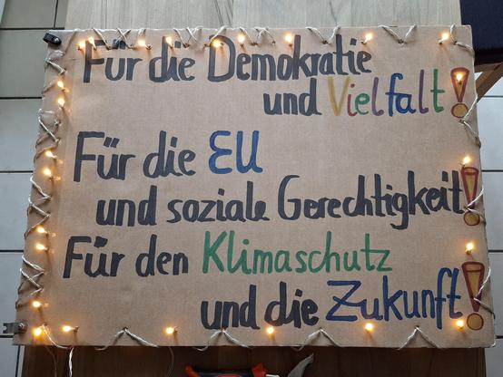 Auf dem Küchentisch liegt ein Plakat. Die Aufschrift:
"Für Demokratie und Vielfalt! 
Für die EU und soziale Gerechtigkeit! 
Für den Klimaschutz und die Zukunft!" Ist jetzt mit einer Lichterkette umrandet, die auch die Rückseite mit dem gleichen Text beleuchtet.