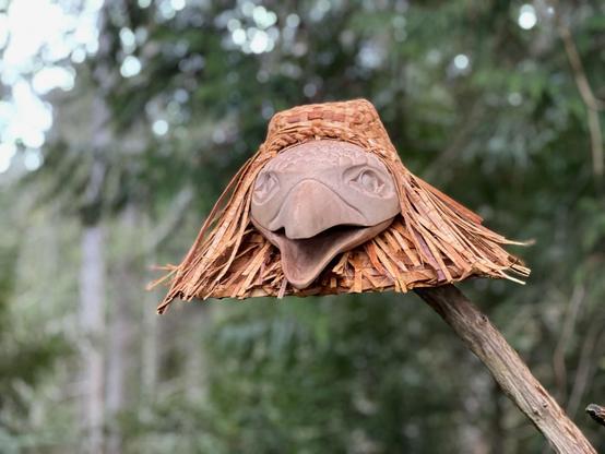 Close up of cedar woven Coast Salish hat adorned with a carved raven's head perched atop a wooden pole, displayed outdoors, with a blurred background of a forest environment. The hat is light brown/beige & densely woven, with cedar strip strands fringed on both sides of the raven head face carving. The raven carving is pale pinkish beige, with an open beak & an intricate Coast Salishan eye design.
Front facing the camera.
