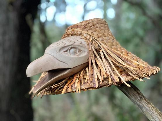 Close up photo of a cedar woven Coast Salish hat adorned with a carved raven's head perched atop a wooden pole, displayed outdoors, with a blurred background of a forest environment. The hat is light brown/beige & densely woven, with cedar strip strands fringed on both sides of the raven head face carving. The raven carving is pale pinkish beige, with an open beak & an intricate Coast Salishan eye design.
Left profile view.