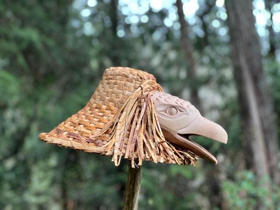 Close up photo of a cedar woven Coast Salish hat adorned with a carved raven's head perched atop a wooden pole, displayed outdoors, with a blurred background of a forest environment. The hat is light brown/beige & densely woven, with cedar strip strands fringed on both sides of the raven head face carving. The raven carving is pale pinkish beige, with an open beak & an intricate Coast Salishan eye design.
Right profile view.