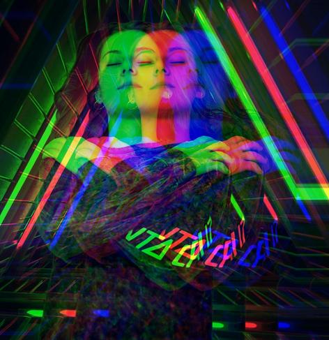 Close-up, medium-shot portrait. Subject's face is triplicated with chromatic aberration effects, using RGB (Red, Green, Blue) channel separation. Woman's eyes are closed. She is wearing a printed top. Background a dark & dimly lit, triangular grid lit with RGB LEDs creating strong diagonal lines. Composition emphasizes layering & optical distortion.