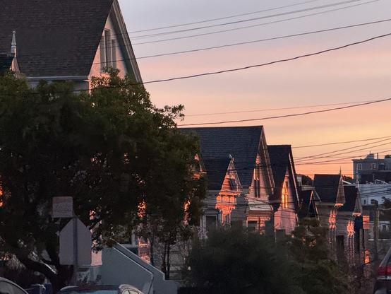 A row of houses with sloped roofs is illuminated by the warm glow of sunrise, framed by trees and power lines.
