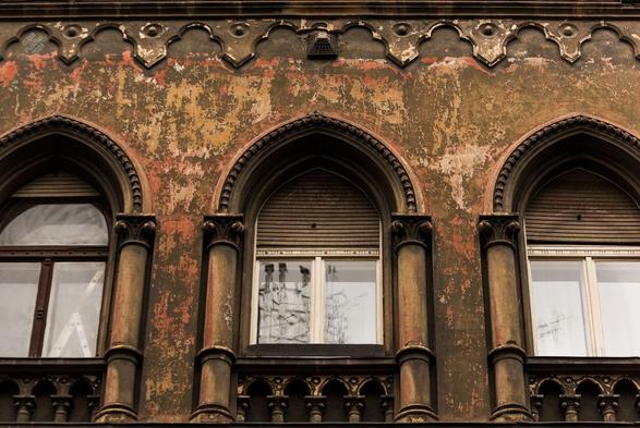 Looking up at a building's second floor. There are three gothic arched windows, with decorative balustrades, columns, and arches, surrounded by a plastered wall that shows the effect of weathering revealing many layers of paint; at the top, a decorative "gothic" cornice.