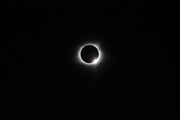 The picture depicts a total solar eclipse. Most of the sky is completely dark. The sun is a dark void surrounded by a white flame that seems to glow without illuminating anything. In the bottom right, the sun is just barely peaking through, appearing as a bright flare.

In the background you can see various stars. They each have a visibly different color, but they are faint enough that you might miss them if you weren't looking. Each star appears with a slight double image, because I was shaking the camera a bit too much