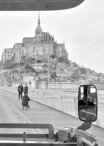 A crouching person photographs two people standing in front of them. In the background is Mont Saint-Michel. In the foreground, a rear-view mirror reflects two children.
***
Une personne accroupie photographie deux personnes debout devant elle. En arrière-plan il y a le Mont Saint-Michel. Au premier plan, un rétroviseur reflète deux enfants.