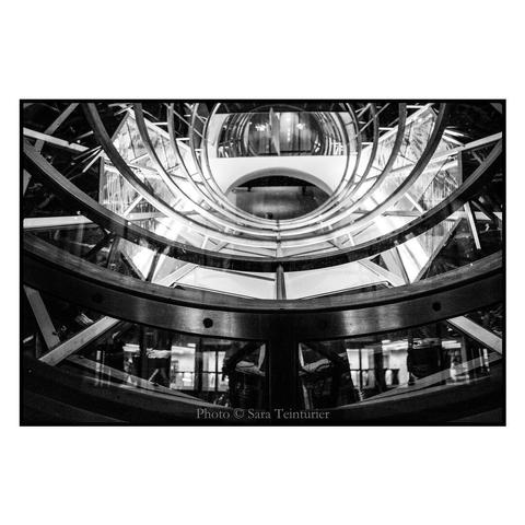 (English follows) Photographie noir et blanc, format paysage, ratio 3x2. L'image représente le détail d'un puit de lumière circulaire, en métal et verre. Elle met en valeur la symétrie de sa conception, et sa relative complexité : le nombre de "bras" de métal est important. Dans le verre, en bas, on devine des silhouettes. Hors de tout contexte, il est difficile d'imaginer ce que représente l'image, lui conférant  une dimension d'abstraction non négligeable. 

Black and white photograph, landscape format, 3x2 ratio. The image shows the detail of a circular metal and glass skylight. It highlights the symmetry of its design, and its relative complexity: the number of metal “arms” is significant. Silhouettes can be made out in the glass below. Taken out of context, it's hard to imagine what the image represents, lending it a certain dimension of abstraction.