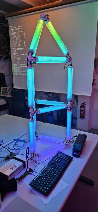 Bunte Lightpipes stehen auf einem Tisch. Dahinter hängt eine Leinwand, auf dem Code zu sehen ist.