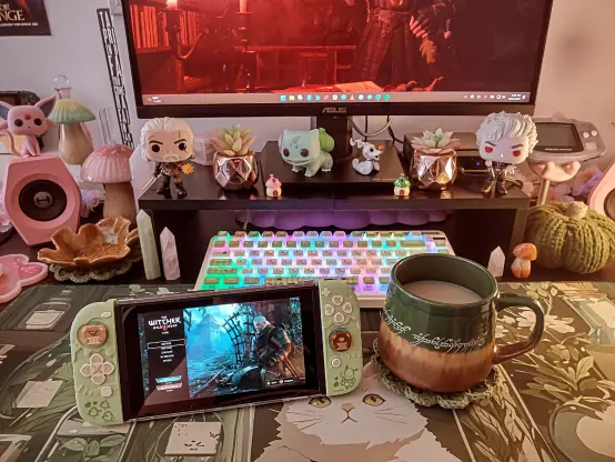 This is a photo of my gaming desk with sage and darker green shades, along with pops of pink. I have my Nintendo Switch with The Witcher's startup screen on it. It's sitting on my desk and resting next to it is my tea mug, also dark green. On it, it has the inscription on the One Ring from Tolkien's The Lord of the Rings. I have Bulbasaur, Geralt, and Astarion Funko Pops beside my monitor, mushrooms everywhere, and crystals all around.
