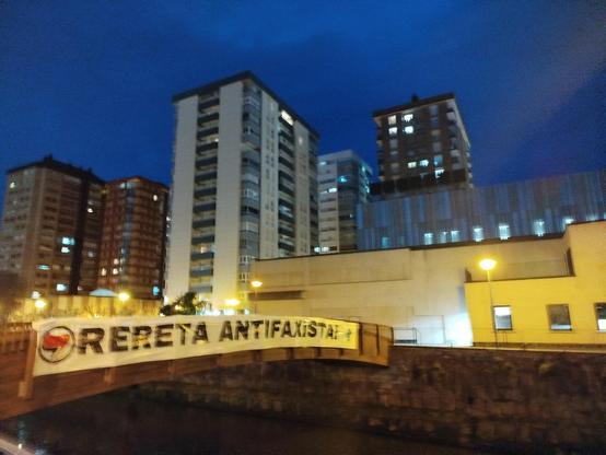 Oiartzun ibaiaren gaineko zubian, Lekuona kultur fabrikaren aurrean zubi guztia hartzen duen pankarta, 'Orereta Antifaxista jartzen du, O-an sinbolo antifaxista bandera gorri eta beltzarekin. Atzean Olibeteko dorreak, zerua ilun.