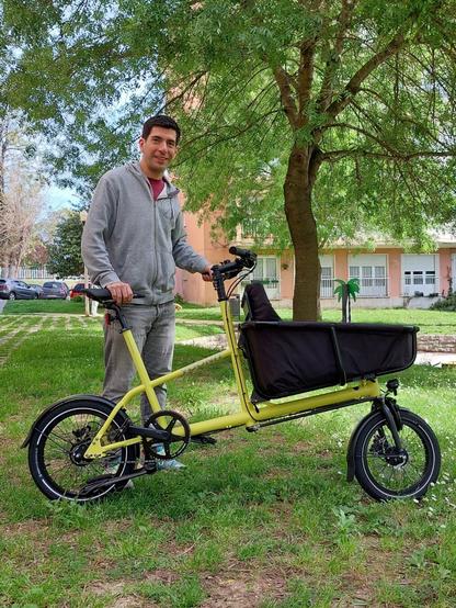 smiling person stands on a grassy lawn, with trees, a building, and cats in the background. The person is standing beside a front-loading cargo bike similar to an Urban arrow but based on a mini-velo platform making the overall length much shorter while managing a similar load capacity.