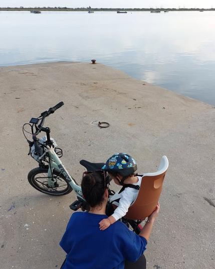 Mãe com criança pequena sentada numa cadeira castanha em cima de uma bicicleta verde junto ao rio