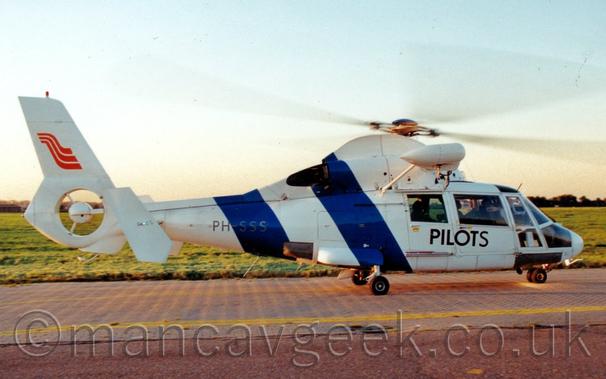Side view of a white helicopter ground-taxiing from left to right.
There are 2 thick, dark blue diagonal  on the rear fuselage and tail boom, and black "Pilots" titles on the side of the passenger cabin doors.
There is a red, 3-part wave on the tail, above the shrouded tail rotor.
Grey taxiway fills the foreground, with grass filling the background, leading up to trees in the distance, under bright but hazy skies.