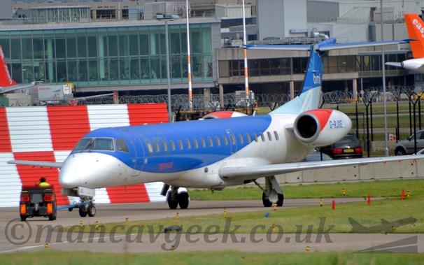 Side view of a white and blue, twin engined jet airliner with it's engines mounted on the sides of the rear fuselage, facing to the left but being pushed back to the right by a small grey tug.
The plane has a white belly, and blue upper fuselage.
The tail has a light blue lower half and dark blue upper half, with white "BMI" titles on the top section.
The top of the engine pod is red at the front and white everywhere else, with the registration "G-RJXE" in blue in the middle.
Green grass fills most of the foreground, lining the edges of taxiways, with a red and white checkerboard metal frame in the background on the left of the frame.
A large grey terminal building stretches across the rest of the frame.
