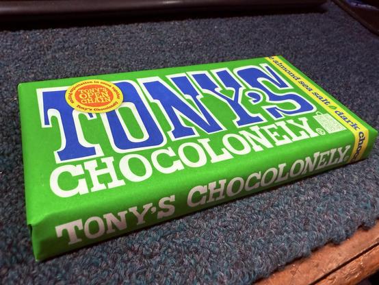 This is an image of a Tony's Chocolonely chocolate bar. The packaging is green, and the text on it indicates it's a dark chocolate with almond sea salt flavor. Tony's Chocolonely is known for its commitment to fair trade practices.