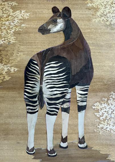 Photograph of a marquetry okapi set in a background. The okapi’s butt is towards the viewer, showing off its stripey pajamas. The wood in the background is a golden color, and there is stylized golden foliage around the okapi.