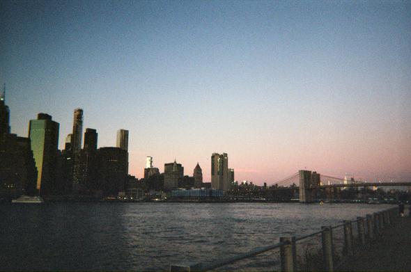 A dark, grainy film cityscape. The Manhattan skyline in black silhouette along the East River taken from a pier in Brooklyn. The daylight is rapidly fading - just a pink strip of sunset remains below a dark blue sky.