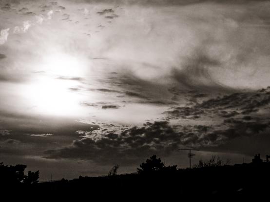 This is a black and white photograph capturing a dramatic sky filled with dense, layered clouds. The sun is hidden behind the clouds, creating a bright, glowing area in the upper left portion of the image. The clouds are thick and textured, with varying shades of gray, giving a sense of depth and movement. The lower part of the image features silhouettes of trees and rooftops, suggesting an urban or suburban setting. The overall mood is atmospheric and somewhat moody, highlighting the contrast between the bright sky and the darker foreground elements.