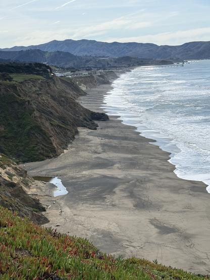 Coastal view featuring sandy shorelines, distant cliffs, and gentle waves. The landscape includes green vegetation in the foreground, hills in the background, and a pier extending into the ocean.