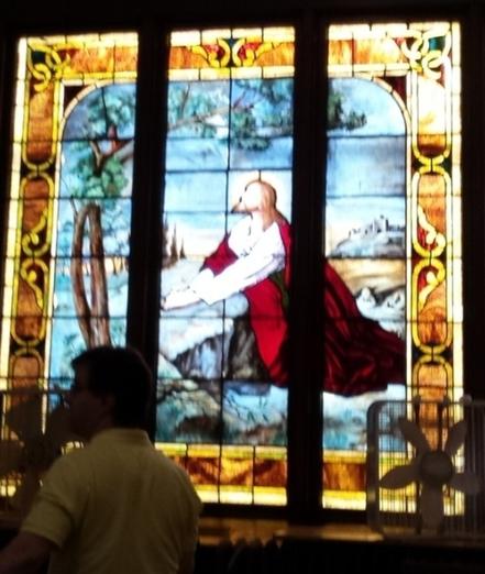 A close-up view of the stained glass window. The window depicts Jesus, clad in white with a red robe, kneeling down and praying by a large rock, and looking heavenward. 

In the foreground, a corner of the window is obscured by the shadow of a man walking past, and a box fan is sitting in the windowsill by the other corner.