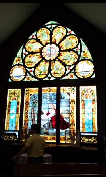 A full view of the stained glass window at the United Methodist Church in Plano, Illinois.

The upper section of the window is a slightly pointed arch shape with a flowery design in the middle. The petals of the flower contain identical jewel images. A small circle is on each side of the flower, but unfortunately, the photo is not high-quality enough to determine what the circles are supposed to be. 

The main section of the window depicts Jesus, clad in white with a red robe, kneeling down and praying by a large rock, and looking heavenward. 

On each side of the main panel is a tall rectangular side panel with identical blue-themed sky scenes. The bottom of each panel is opened a bit inwards for fresh air. 

In the foreground, a corner of the main panel is obscured by the shadow of a man walking past, and a box fan is sitting in the windowsill by the other corner.