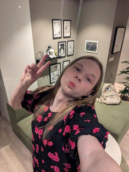 A woman in their mid-twenties, poses for a selfie in her apartment.

Behind them, a shelf displays various figurines and framed artwork.