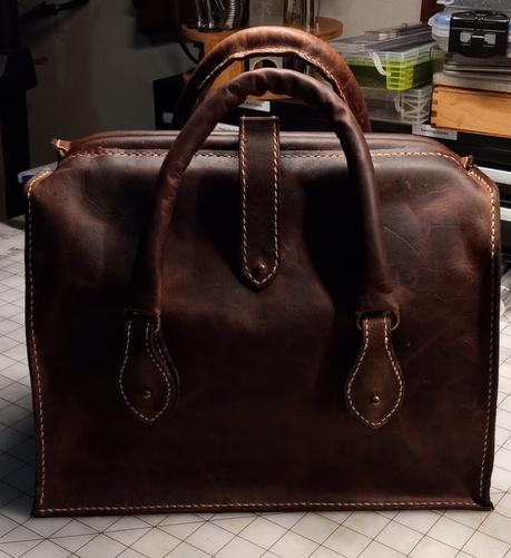 A dark brown leather gladstone style bag. It is about nine inches high, eight inches wide and twelve inches long. It is made from pull up leather that is treated with wax and oils to give it variable tones and make it last longer. It has two tabs on the front and two tabs on the back that hold simple rounded leather handles. There is a strap that comes over the top of the bag to hold it closed, though this is not strictly necessary because the frame of the bag holds it closed. The strap is secured with a Chicago screw.