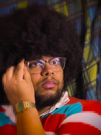 L'image est un portrait d'une personne (moi-même) aux cheveux crépus portant des lunettes, un anneau dans le nez et une chemise à rayures rouges, turquoises et blanches. Son regard est porté au loin, orienté à 14H. Elle porte une barbe et une moustache et pince la branche droite de ses lunettes avec sa main gauche portant une montre. L'arrière-plan est constitué d'un tissu à motifs quadrillés en différentes nuances de verts et de bleus.