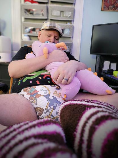 A middle-aged man, wearing a t-shirt, fuzzy socks, and a thick, cartoon printed adult diaper, hugs a plush dragon close while leaning back in his desk chair.