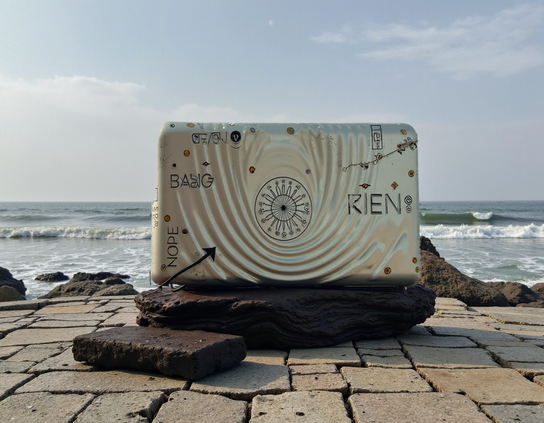 A surreal metallic suitcase-like object engraved with nihilistic symbols and text, resting on rough stone pavers, by a calm ocean  under a pale blue sky.