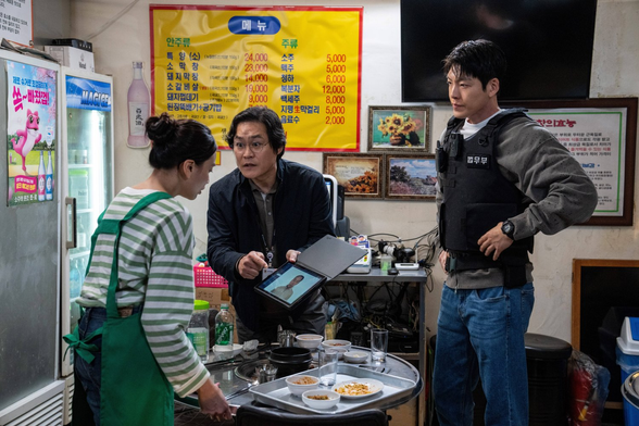 Kim Sung Kyun & Kim Woo Bin dressed as police officers, showing an image on a tablet to a server in a scene from the 2024 South Korean movie, "Officer Black Belt."