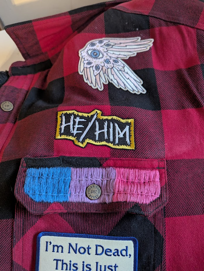 A "He/Him" pronoun patch with a gold border on a buffalo check battle jacket.