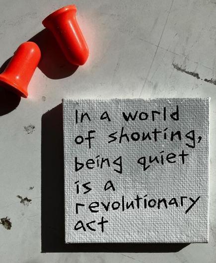 Photography and poetry. The photo shows a small, white, square tablet lying on a table. On it is a handwritten sentence in black ink: "In a world of shouting, being quiet is a revolutionary act". Above it are two orange earplugs.
