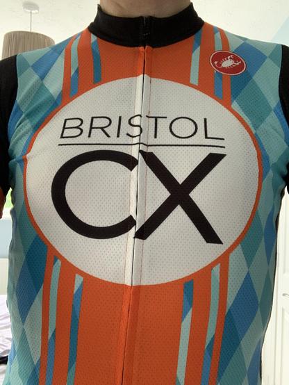 The torso of someone wearing the club jersey of Bristol CX. In the centre is a large white circle with 'BRISTOL CX' written in black in the centre. Running vertically behind the circle are five orange bars. The rest of the jersey consists of a diamond pattern, each diamond in one of several shades of blue. This pattern is called digital camo by the manufacturer. Some kindly say our kit is the best kit in cross.
