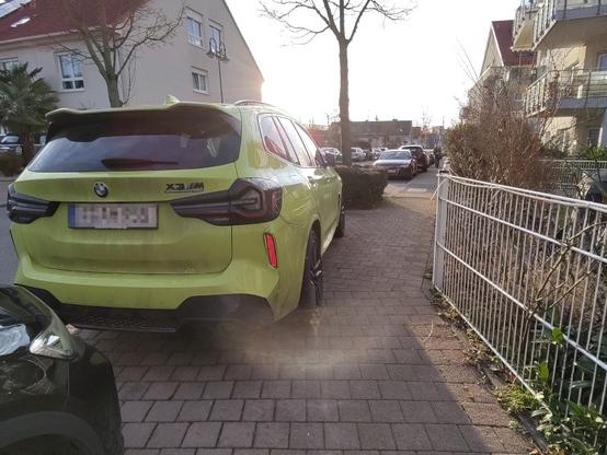 Gehweg, auf dem durch einen Metallzaun rechts und einem falsch geparkten PKW links eine Engstelle gebildet wird. Im Hintergrund Häuser, weitere geparkte PKW und noch kahle Bäume.