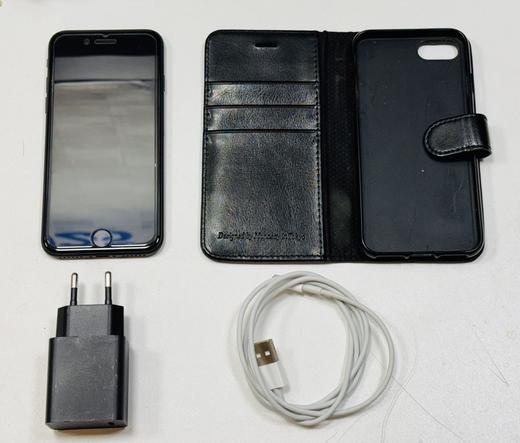 Foto des iPhone SE 2 links oben in der Ecke, darunter das Netzteil in schwarz. Rechts vom Telefon ist eine schwarze Hülle zum Aufklappen und darunter ist ein weißes Ladekabel mit Lightning-Stecker