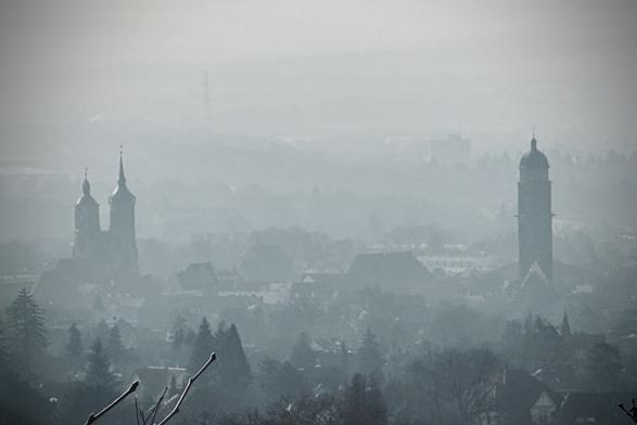 Dunstige Landschaft mit zwei verschiedenen Kirchürmen in gerade so erkennbarer Stadtlandschaft, mit weichen, nebligen Umrissen. Ruhige, atmosphärische Umgebung, typisch für frühen Morgen oder auch Smog.